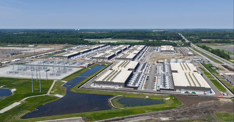 Aerial view of a hyperscale data center campus featuring an adjacent electrical substation and power infrastructure, illustrating the high energy demand required for site selection.